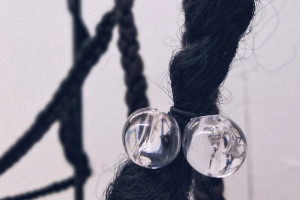 Image is a detail of an installation by artist Ashley Gunter that shows synthetic braided Black hair with hair bobbles wrapped around a wooden frame. The title of the 2019 piece is Holding Space.