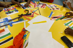 A bright yellow table with colorful art supplies spread across it, showing the hands of Black children sitting around the table creating art. 