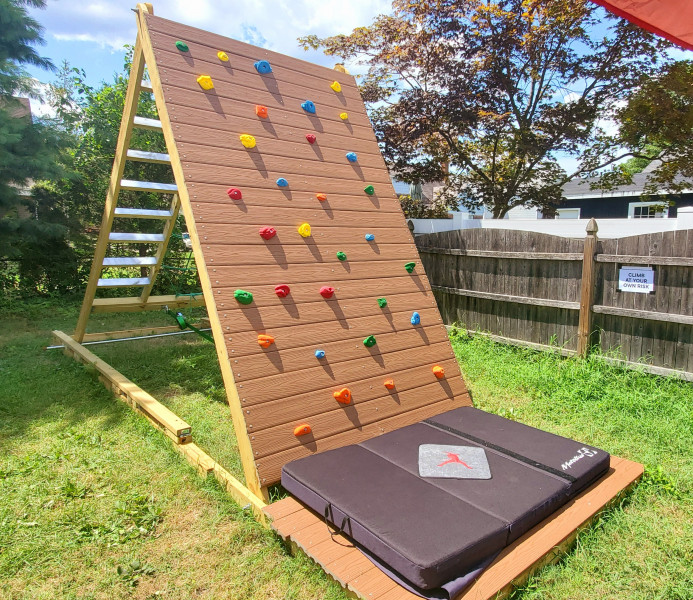 A rock climbing wall made of wood has colorful handles attached to the wall and sits in a grassy green yard.