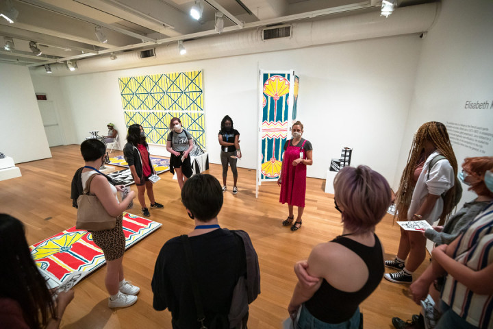 A woman wearing a mask speaking to SADI 2021 students gathered around her in the Fabric Workshop and Museum gallery