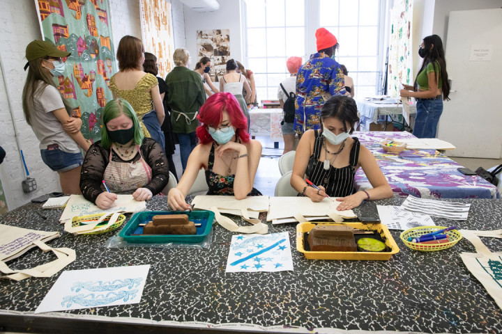 Three SADI 2021 students drawing their designs on tote bags with other students behind them at the Fabric Workshop and Museum