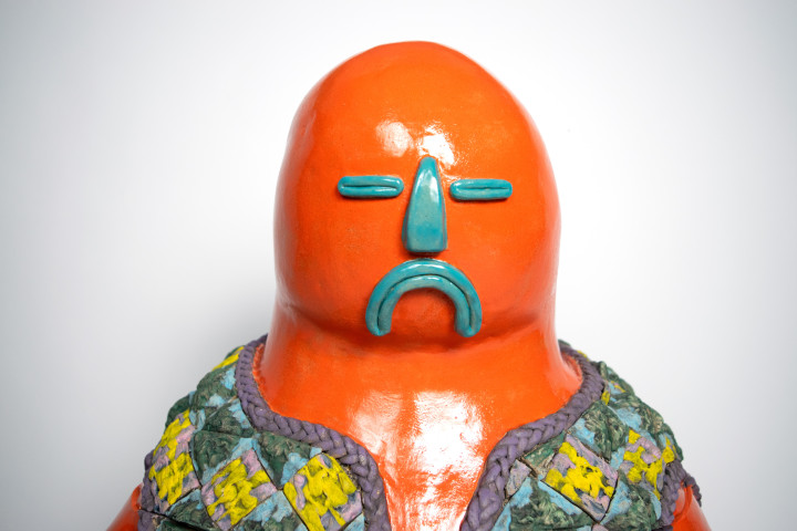 A "Passport-style" photo of a brightly colored sculpture by Hernandez titled "Chiripiorca" with an orange body and turquoise facial features. The shoulders are covered with a woven material. 