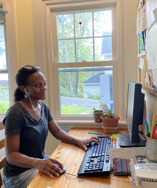 A black women with hair pulled back, her glasses are on top of her head, she has white earphones in her ears, and she is sitting at a table near a window with her computer in front of her.