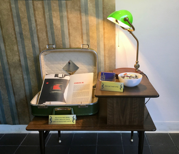 Photo of Christina Faas '21's thesis project. An open publication in an old suitcase on a side table. A lamp, bowl, and business cards are also on the table.